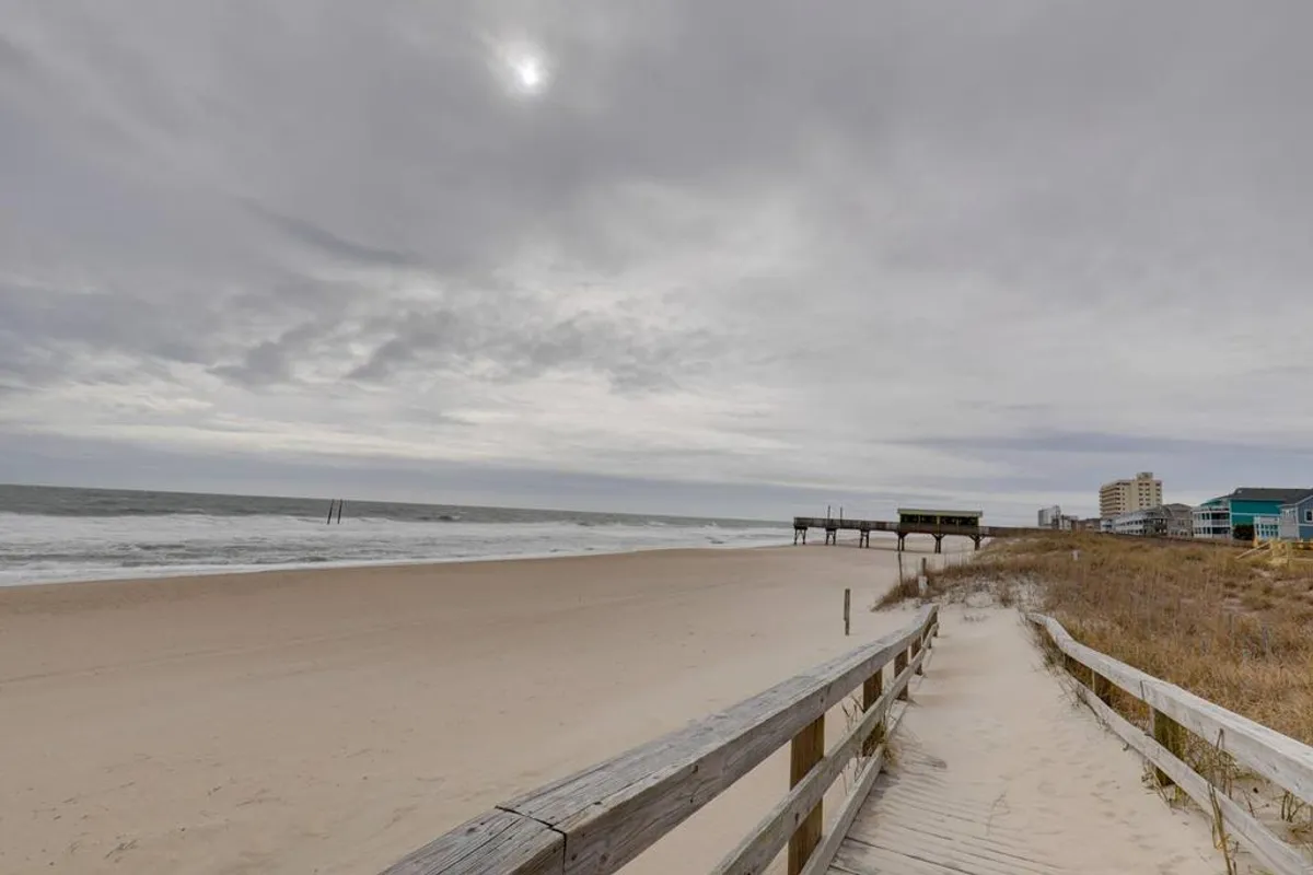 This image shows a boardwalk leading toward the sandy beach, with ocean waves rolling in and a pier visible in the distance under a partly cloudy sky. The inviting path is framed by natural dune grasses, setting a tranquil seaside mood.