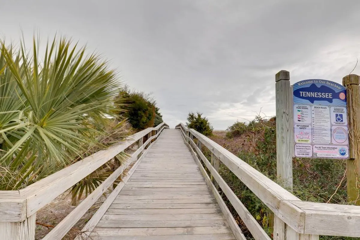 A wooden walkway cuts through lush greenery and palm trees, heading toward the beachfront. An informational sign hints at public access points and the natural beauty that surrounds the approach to the shore.