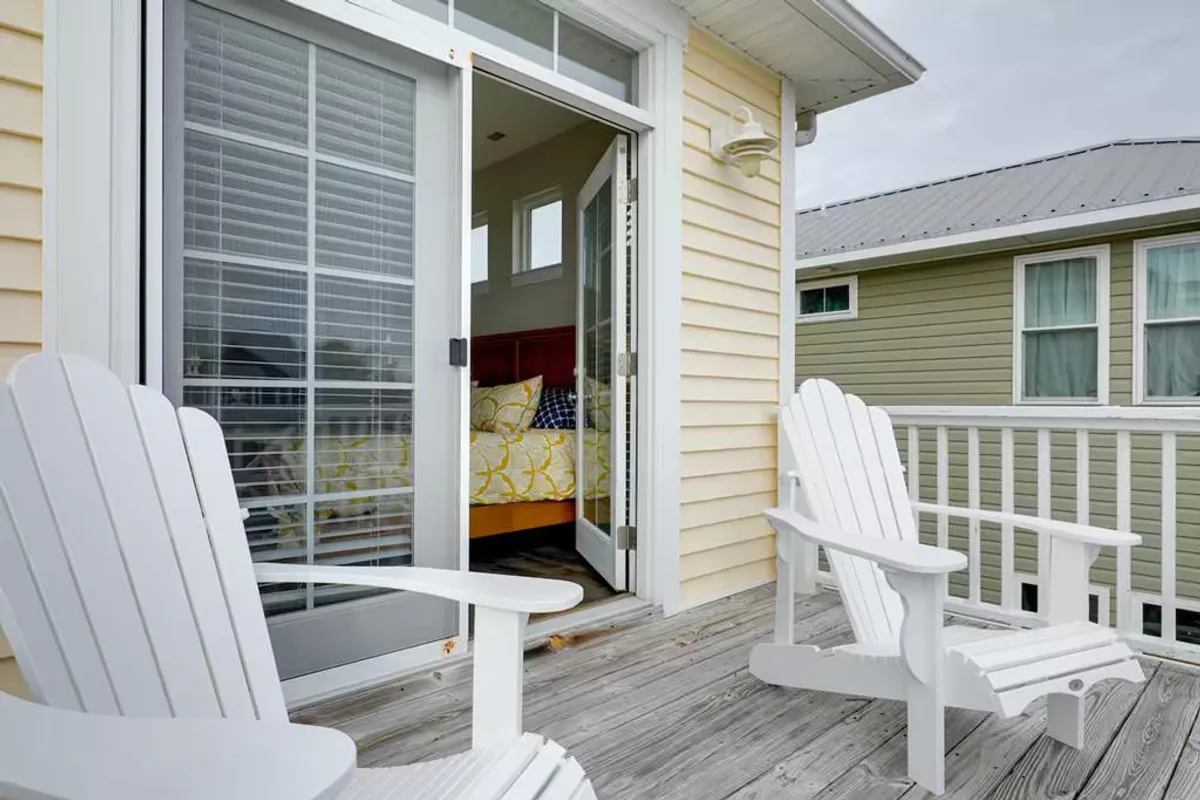 A tranquil bedroom with white Adirondack chairs just outside an open sliding door, and colorful bedding visible inside. The setting encourages unwinding while enjoying the sunset or morning sun from a private deck.