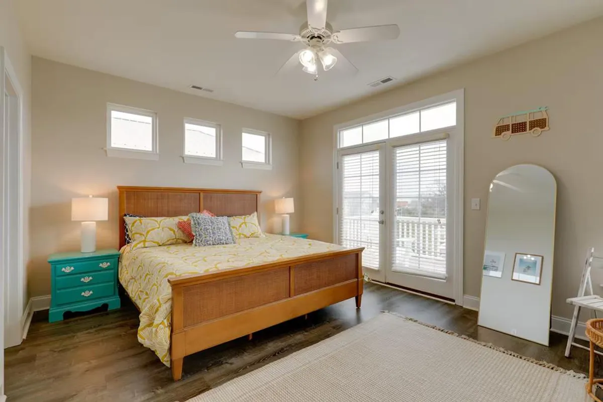 A cheerful bedroom featuring a bed with yellow patterned bedding, a turquoise nightstand, and large windows letting in plenty of natural light. The room feels airy and restful, enhanced by the soft, inviting textures and bright decor.