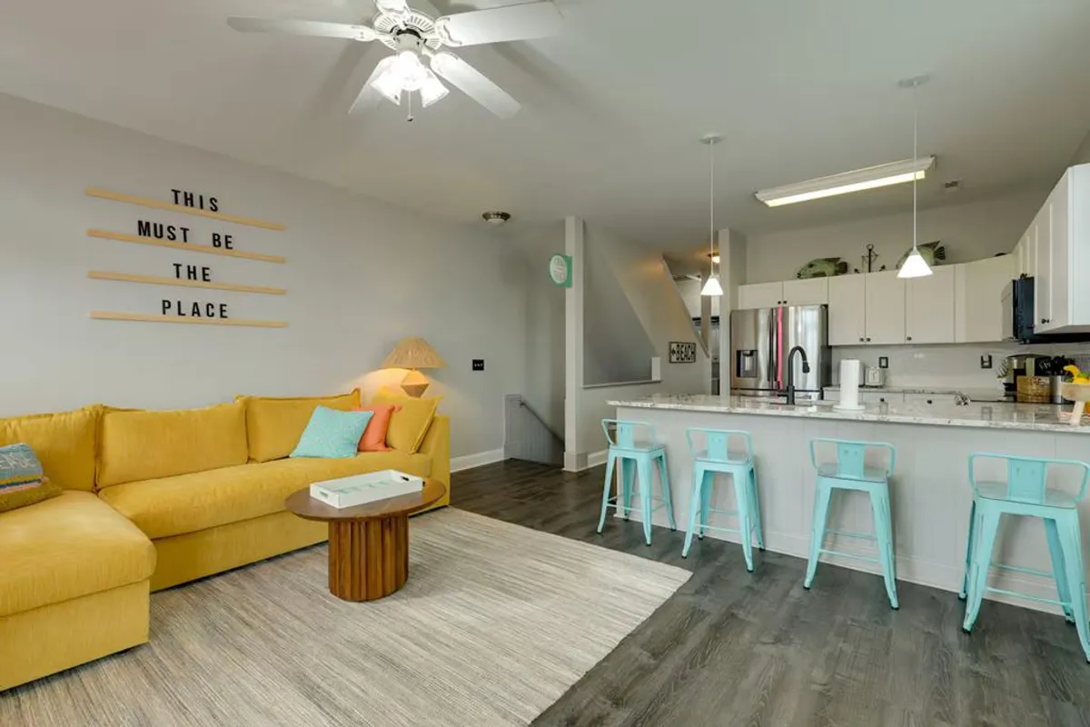 A vibrant, open-concept living area featuring a cheerful yellow sofa, kitchen island with bar seating, and stylish wall art. The scene is bright and inviting, with plenty of space for socializing, casual dining, and enjoying time with friends or family.