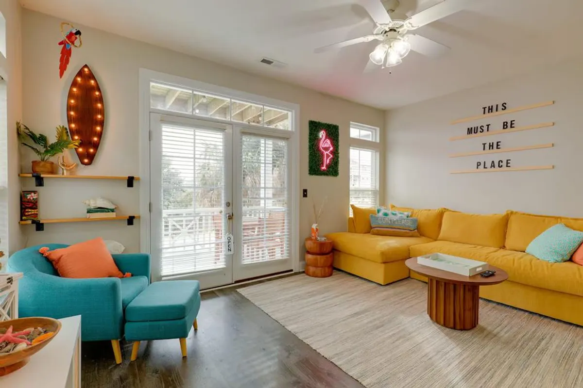 A cheerful living room featuring a bright yellow sectional sofa, playful neon flamingo sign, and colorful accent chair. Decorative shelves and thoughtful details evoke a vibrant, welcoming atmosphere.
